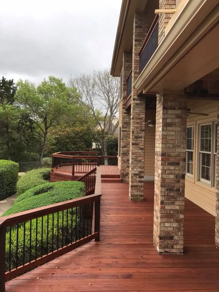 Wraparound stained wood deck along a stone columned two story home with manicured landscaping