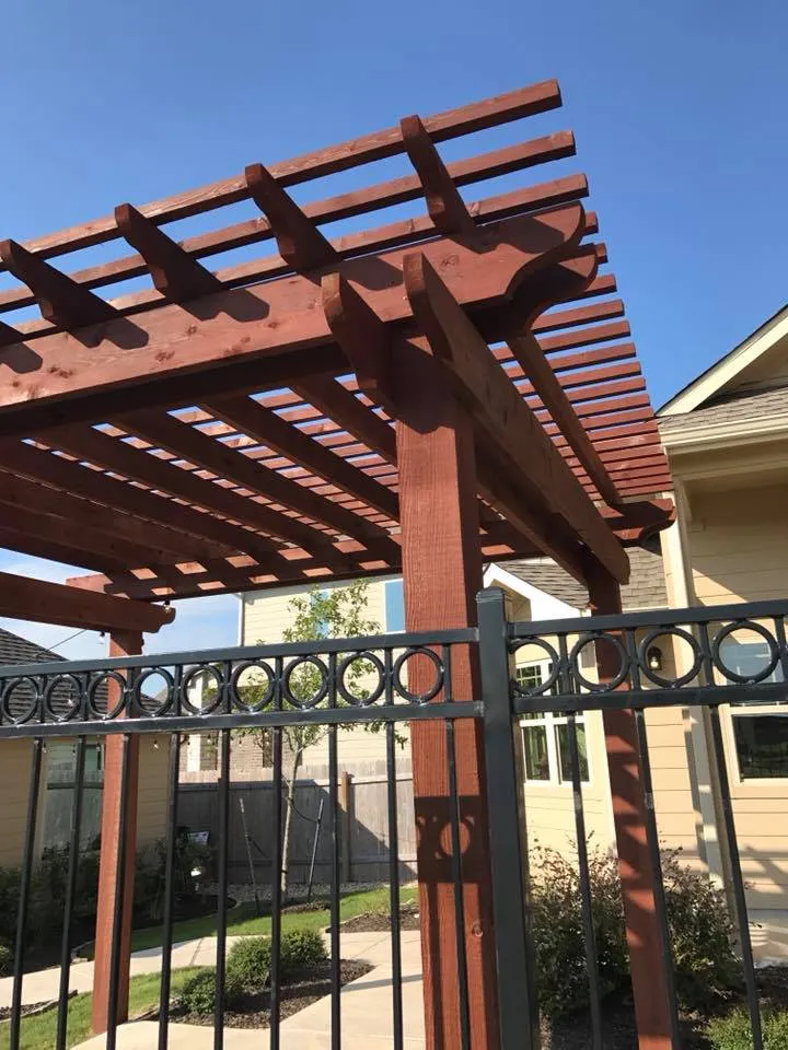 Rich mahogany pergola against blue sky with a decorative black metal fence in front